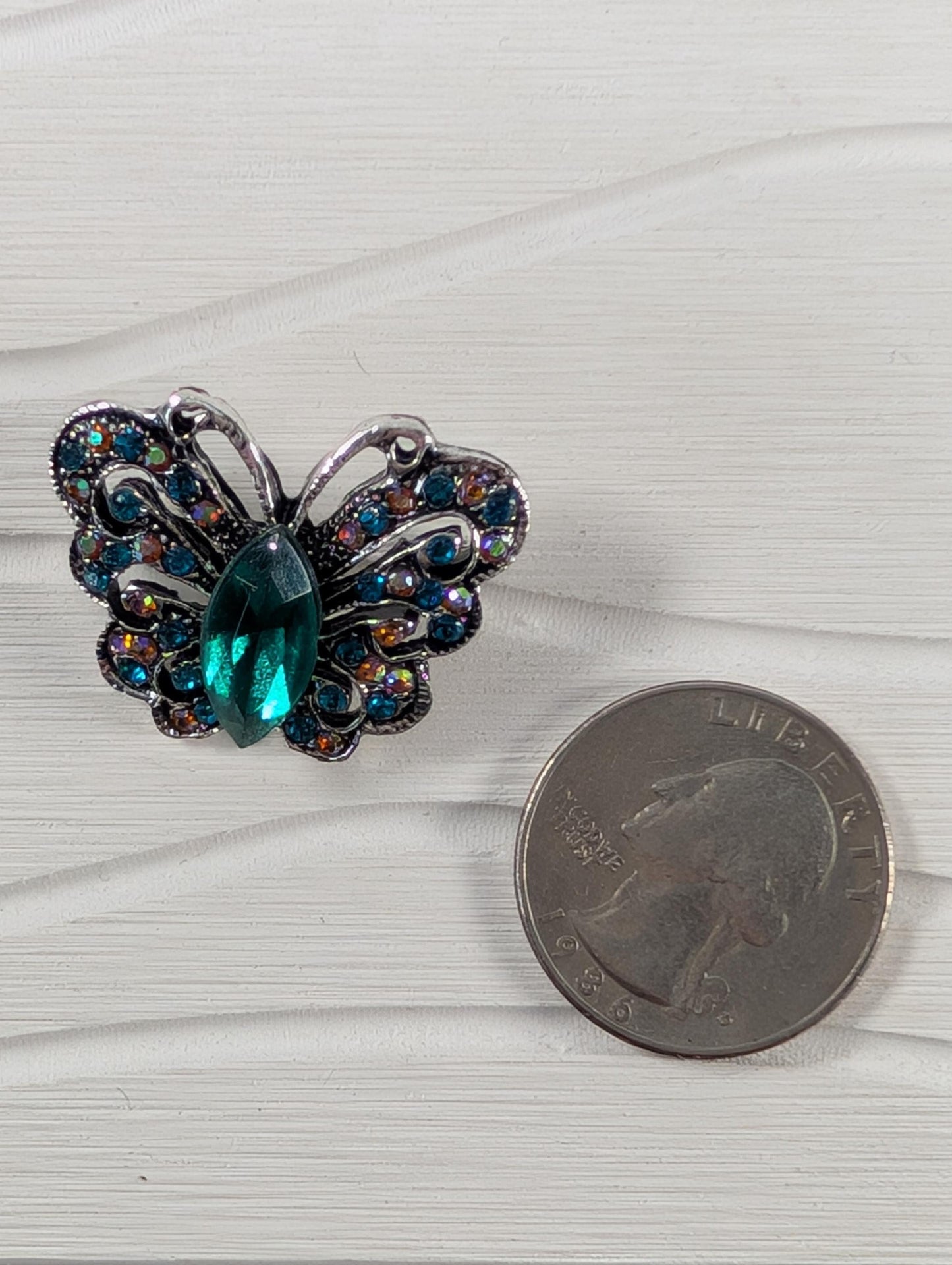 a butterfly-shaped piece of jewelry with a green gemstone on a white wooden surface, next to a United States quarter.