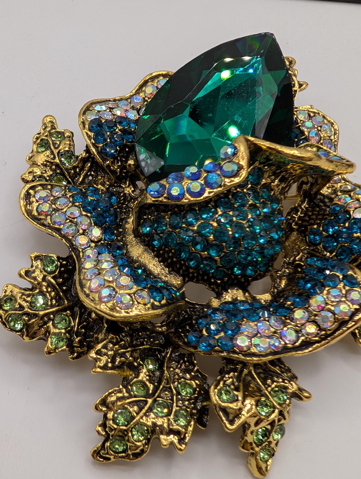 A decorative brooch with a large green gemstone surrounded by smaller blue and green gemstones, all set in an ornate gold and blue floral design.