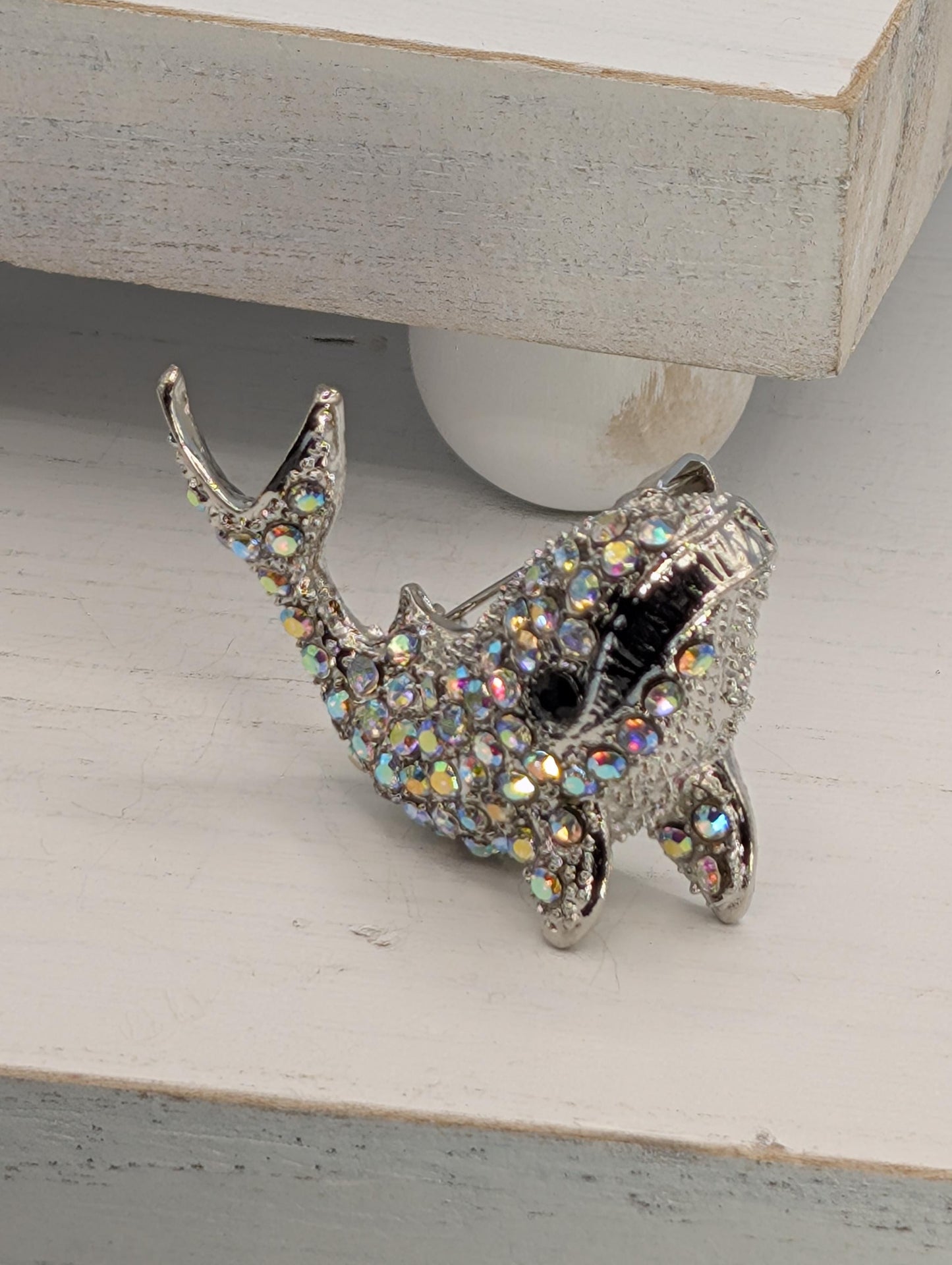 A silver-colored decorative fish brooch with a sparkling crystal design is displayed on a white surface.