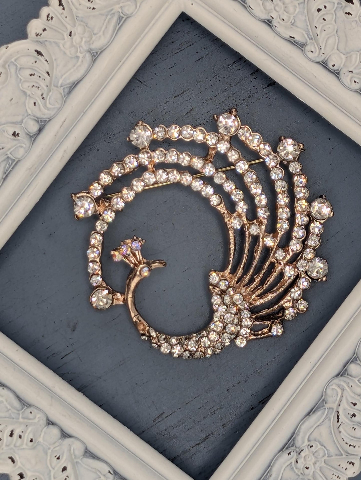 A decorative brooch with a peacock feather design, featuring numerous sparkling rhinestones or crystals arranged in a swirling pattern.