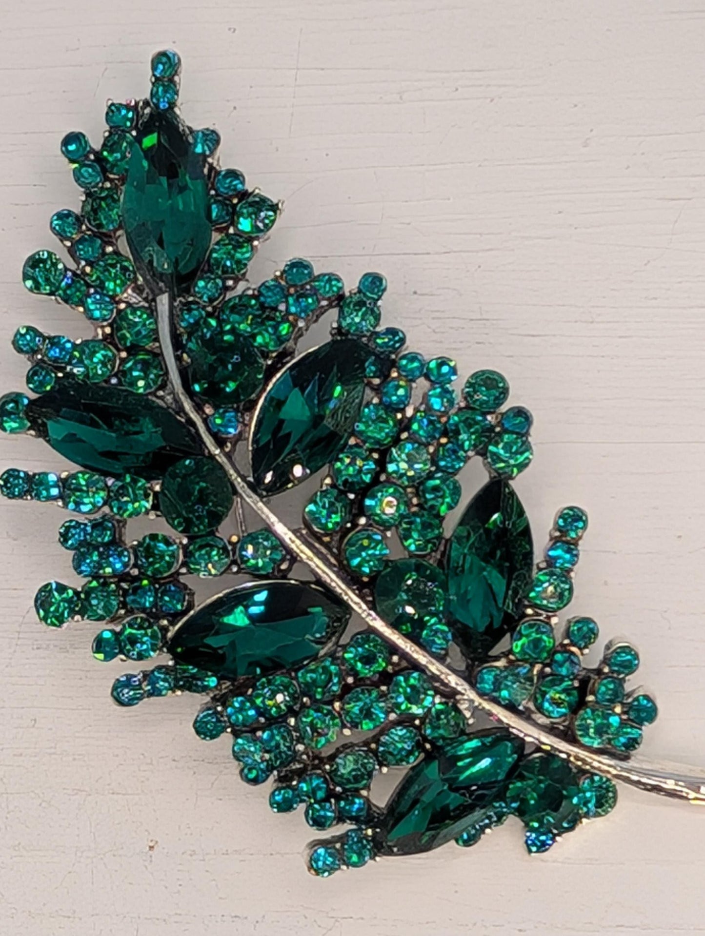 A decorative brooch with a leaf-like design, featuring a variety of green gemstones and metallic elements.