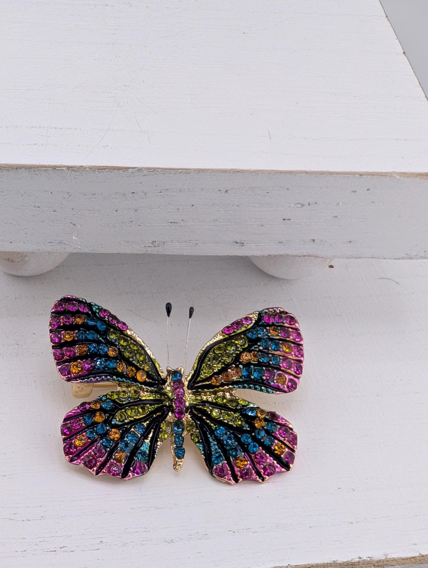 A colorful butterfly brooch with multicolored gemstones is displayed on a white surface.