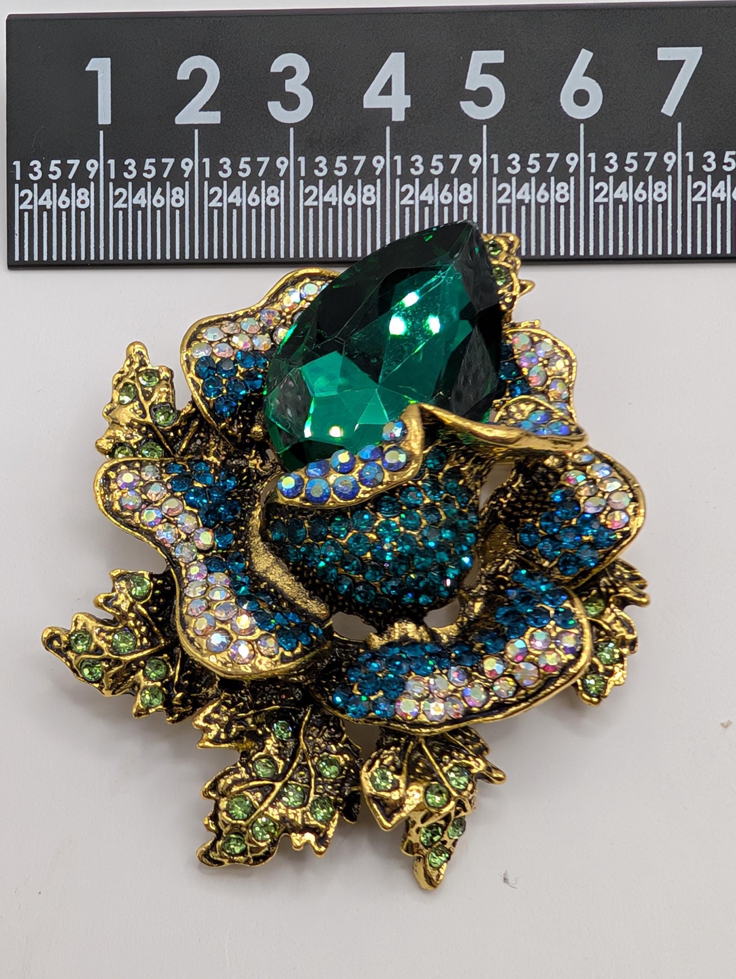 A decorative brooch with a large green gemstone surrounded by smaller blue and green gemstones, set against a black background with a ruler for scale.