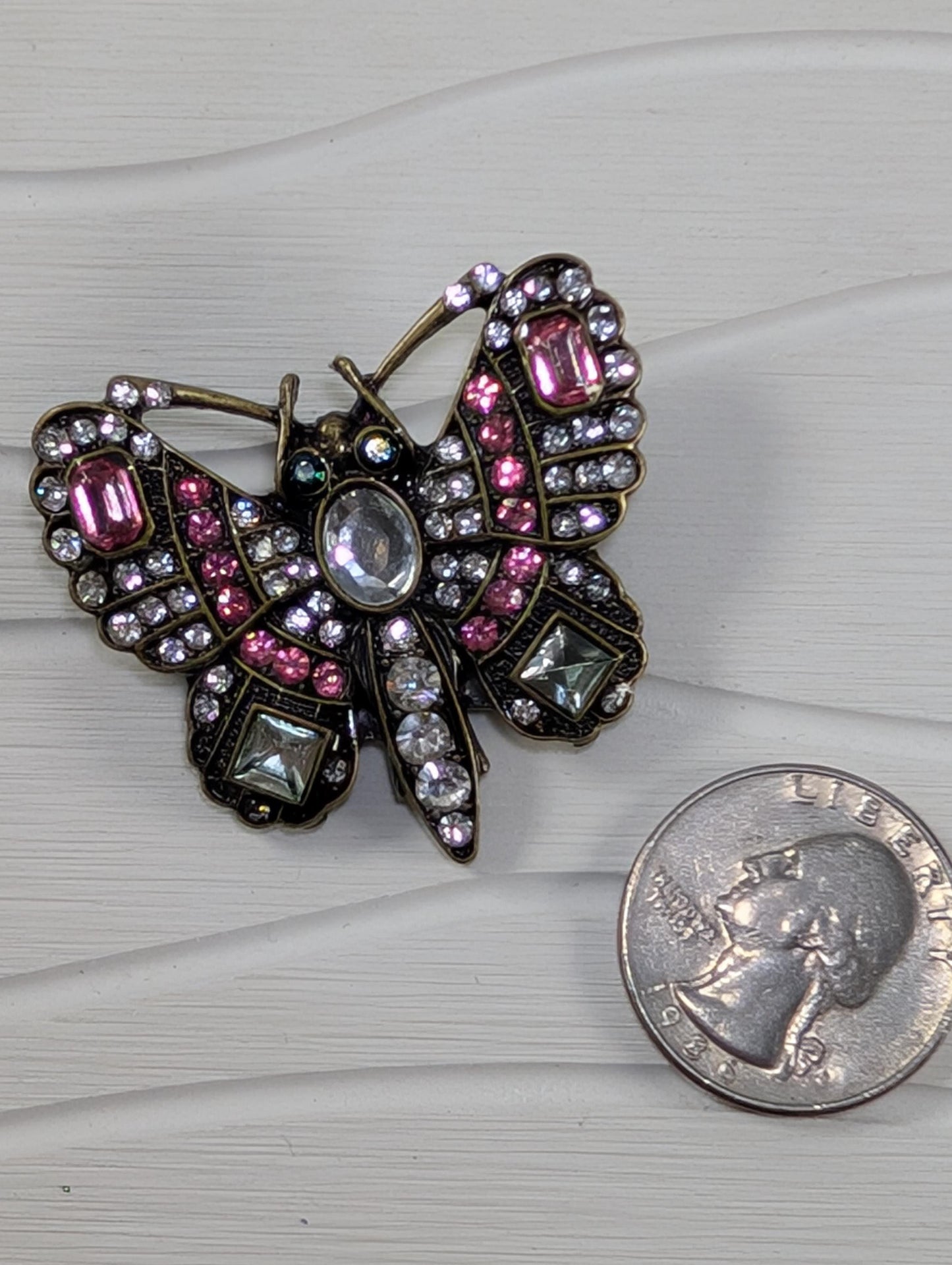 a decorative butterfly brooch adorned with pink and clear gemstones, placed next to a United States quarter.