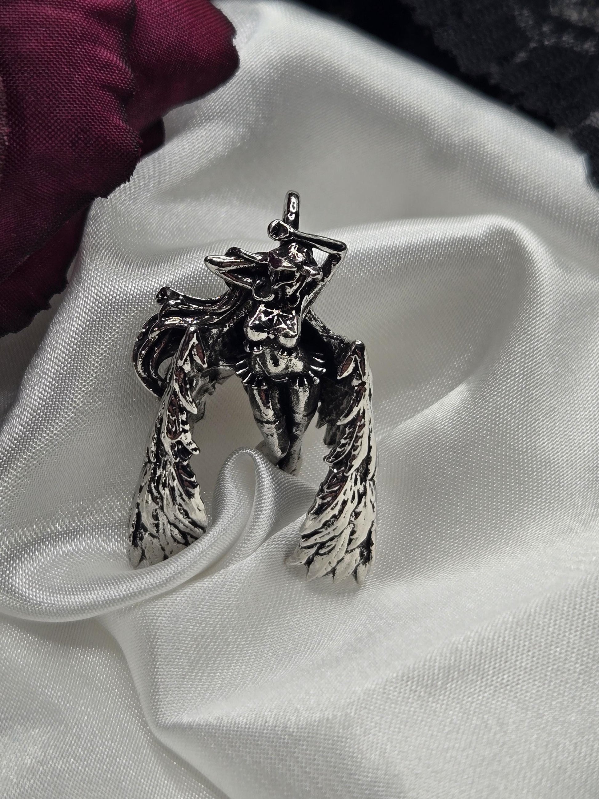 A silver metal winged creature with a skull-like face is displayed on a white fabric surface.