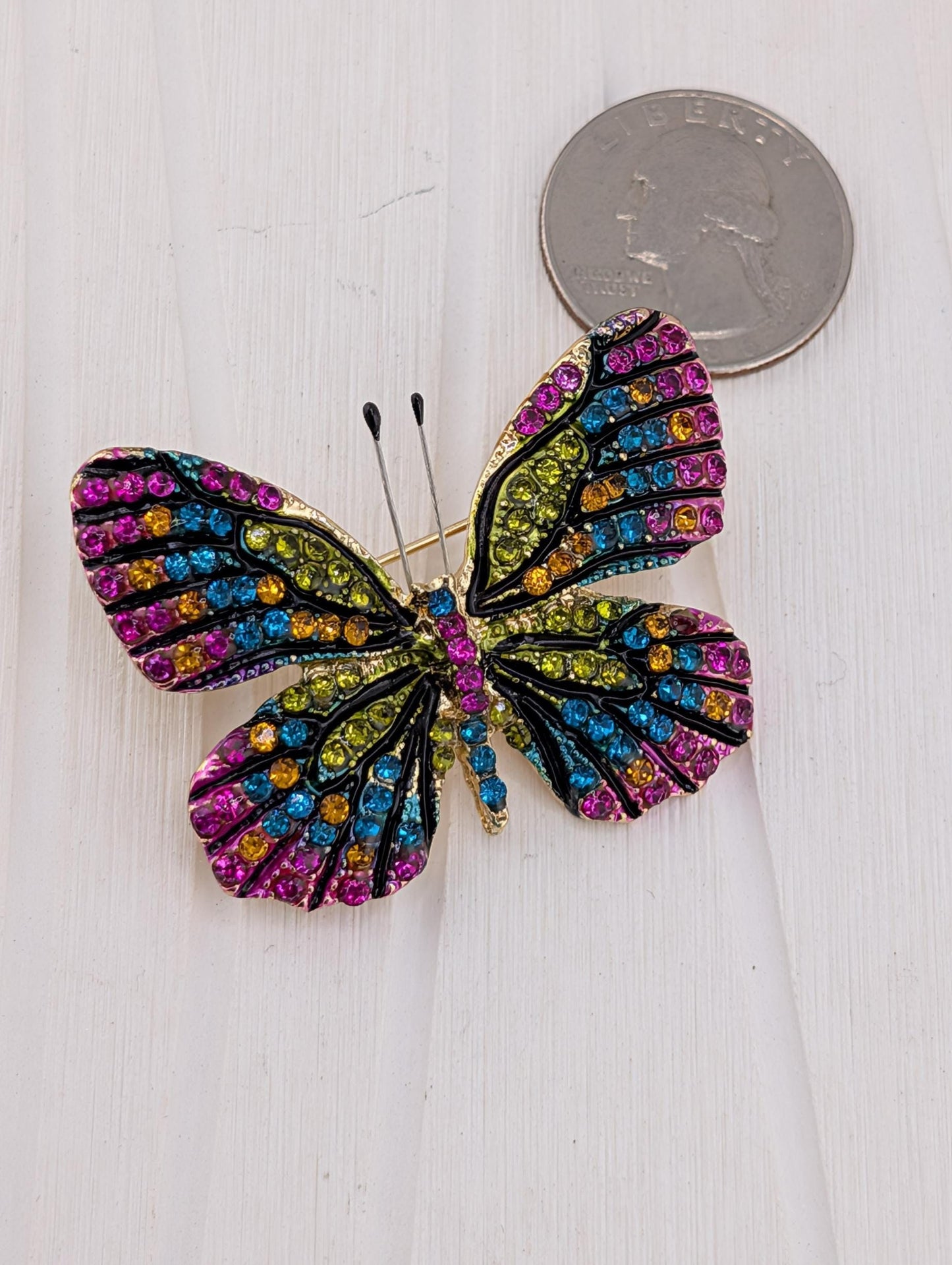 A colorful butterfly brooch with multicolored gemstones is placed on a white surface next to a coin.