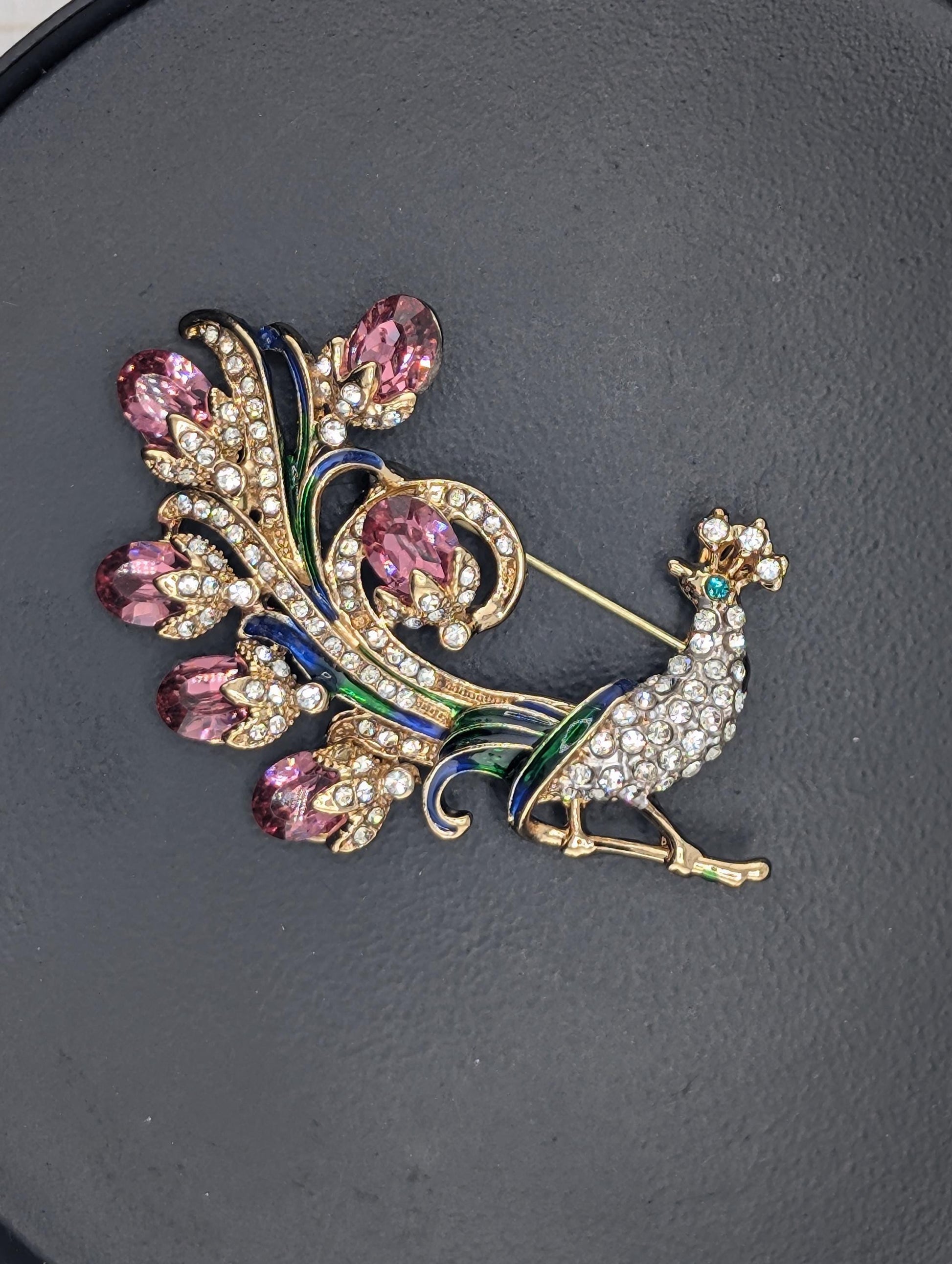 A decorative brooch with a peacock design, featuring a combination of gold, blue, and pink gemstones.