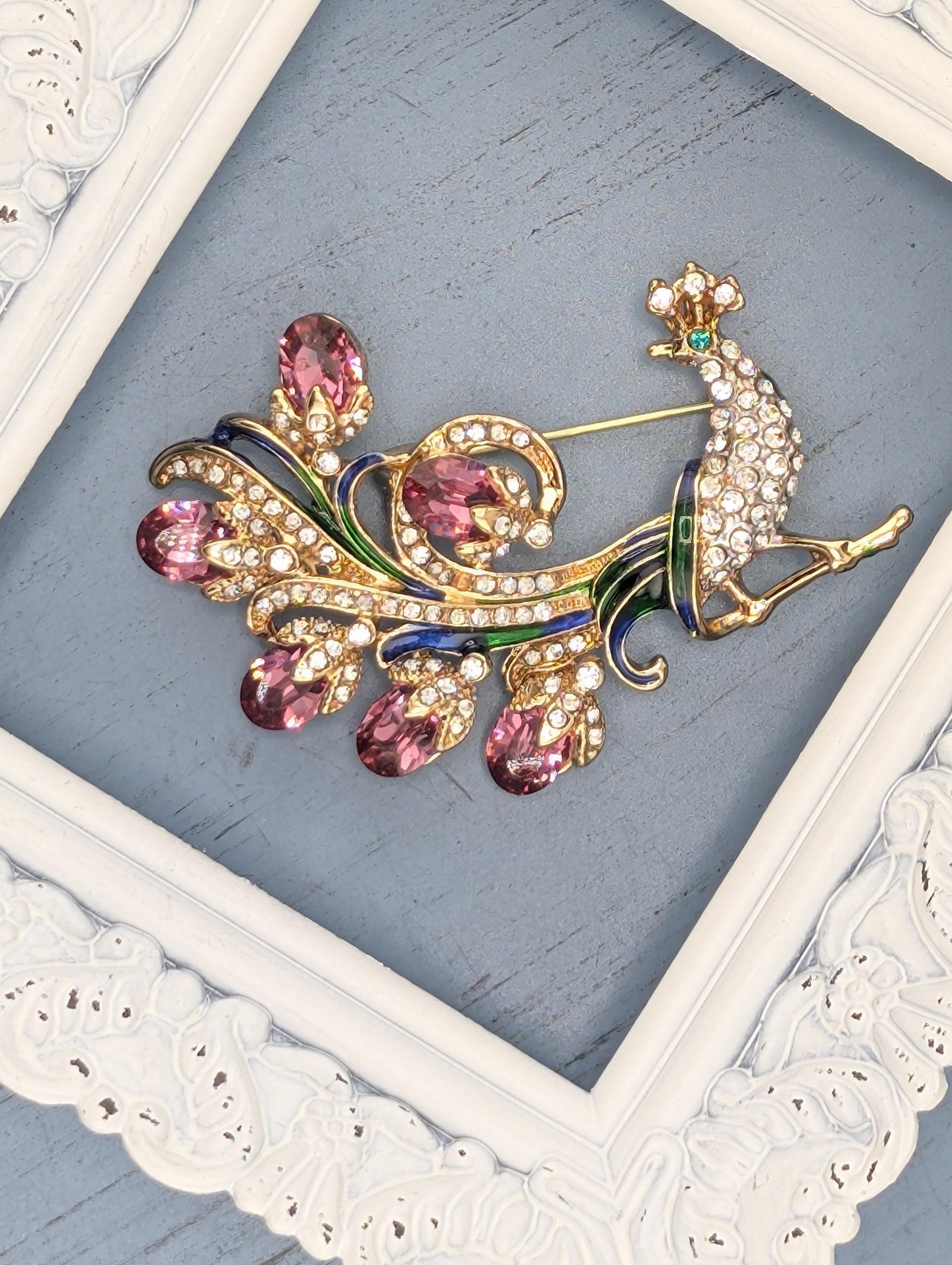 A decorative brooch with a peacock design, featuring a combination of gold, green, and pink gemstones, is displayed on a white ornate frame against a blue background.