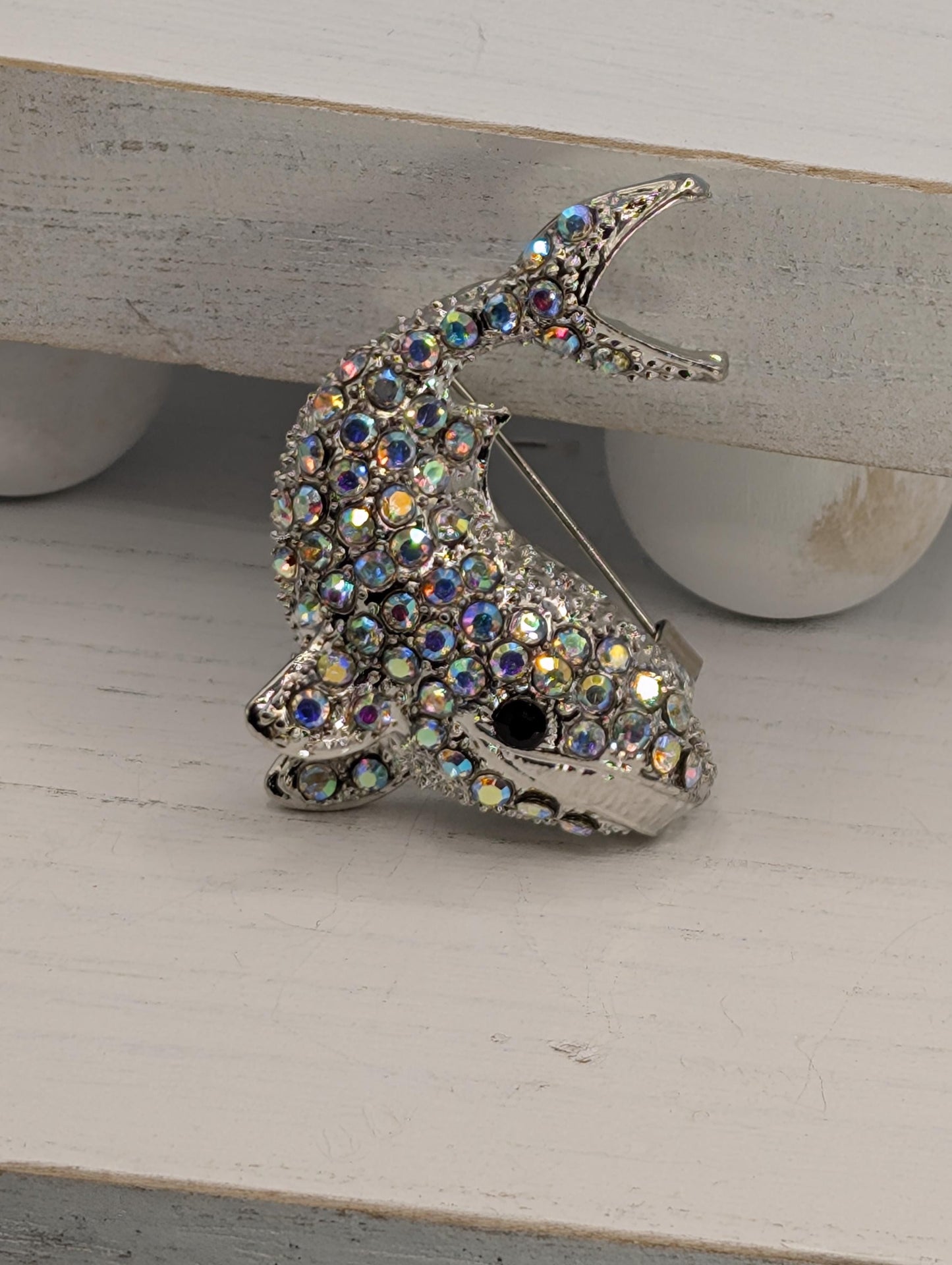 A silver brooch in the shape of a fish, adorned with numerous colorful rhinestones or crystals, is displayed on a white surface.