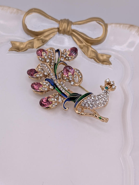 A decorative brooch with a peacock design, featuring a gold-colored bird and a floral pattern with purple and green elements.