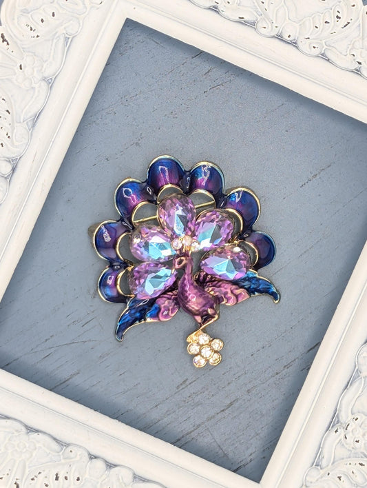 A decorative brooch with a floral design, featuring a combination of purple and blue gemstones, is displayed in a white ornate frame.