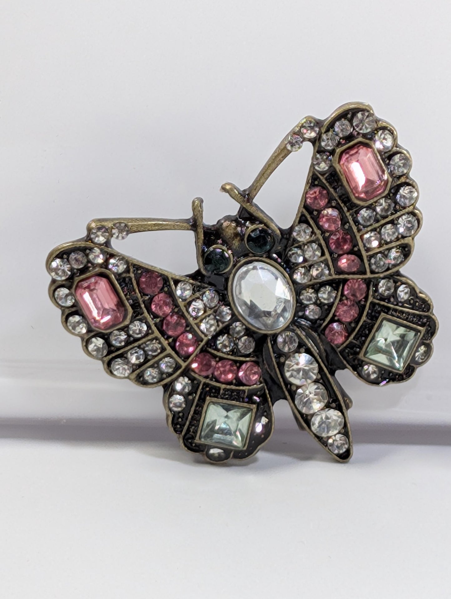 A decorative butterfly brooch with a combination of pink, green, and white gemstones, as well as a clear gemstone in the center.