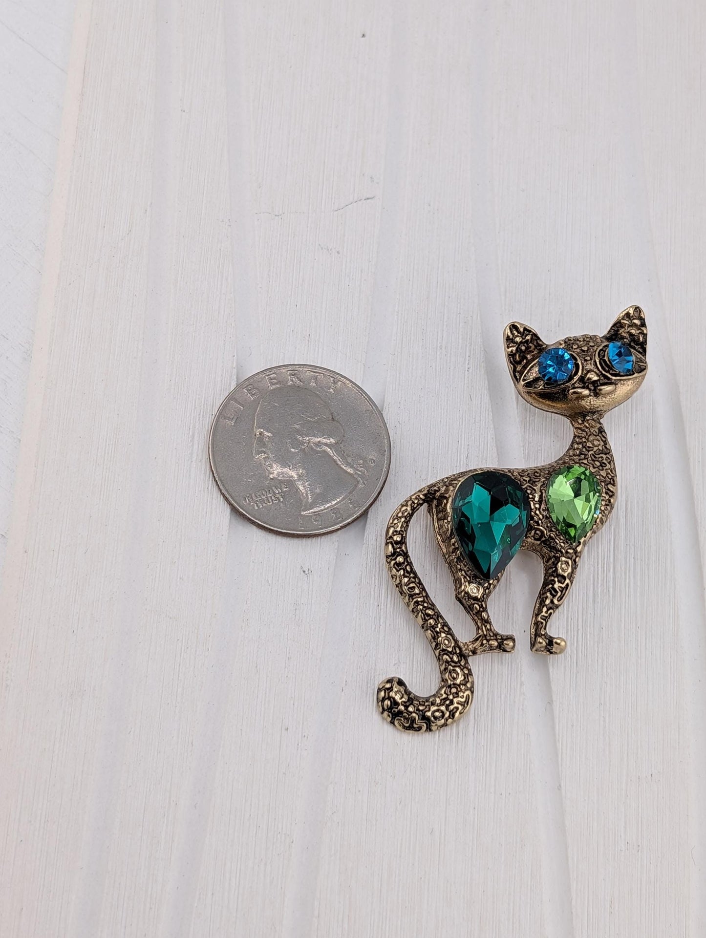 A shiny, ornate cat-shaped brooch made of metal and adorned with colorful gemstones is placed next to a US quarter on a white surface.