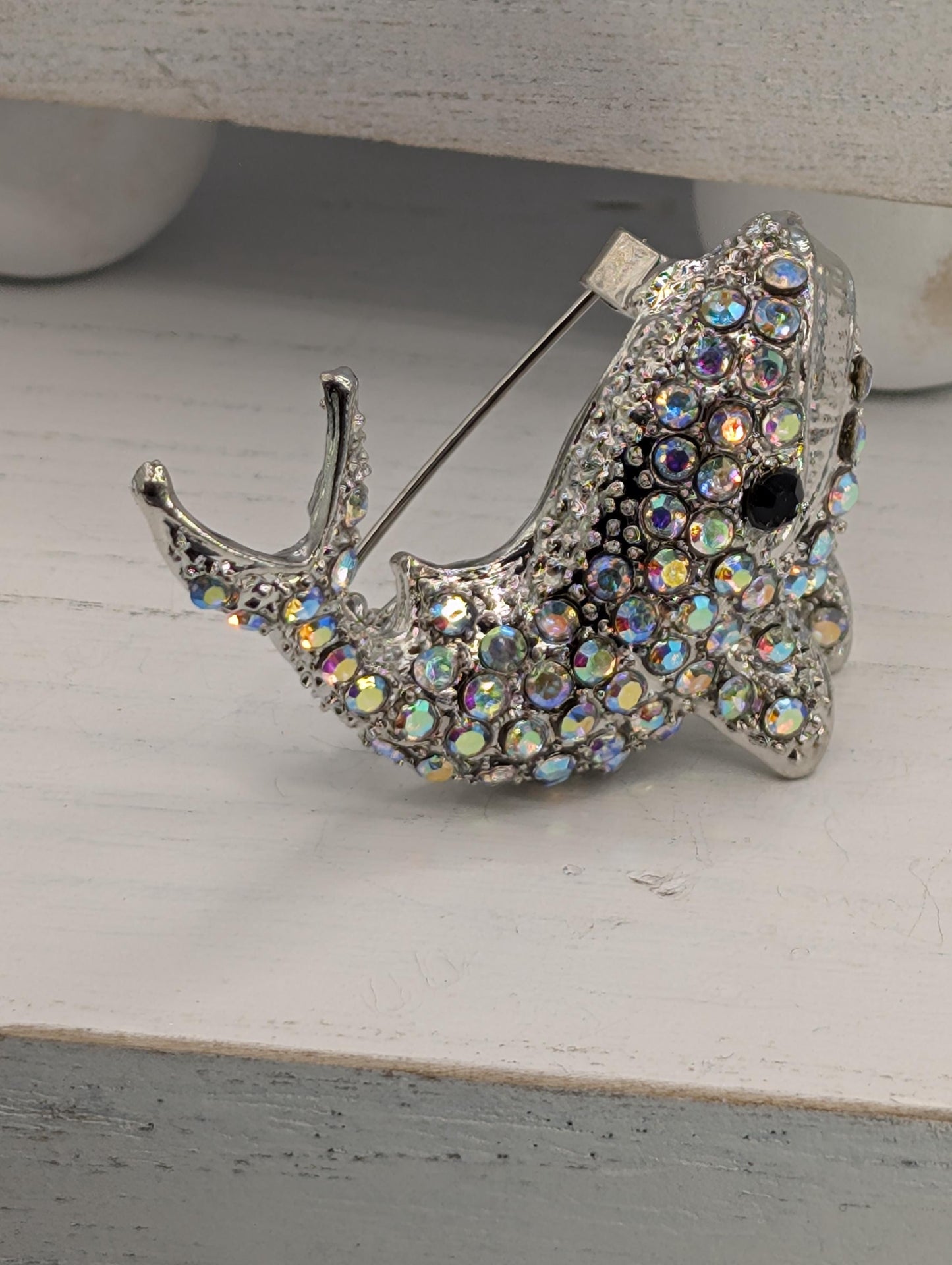 A silver brooch in the shape of a fish, adorned with multicolored rhinestones, is displayed on a white surface.