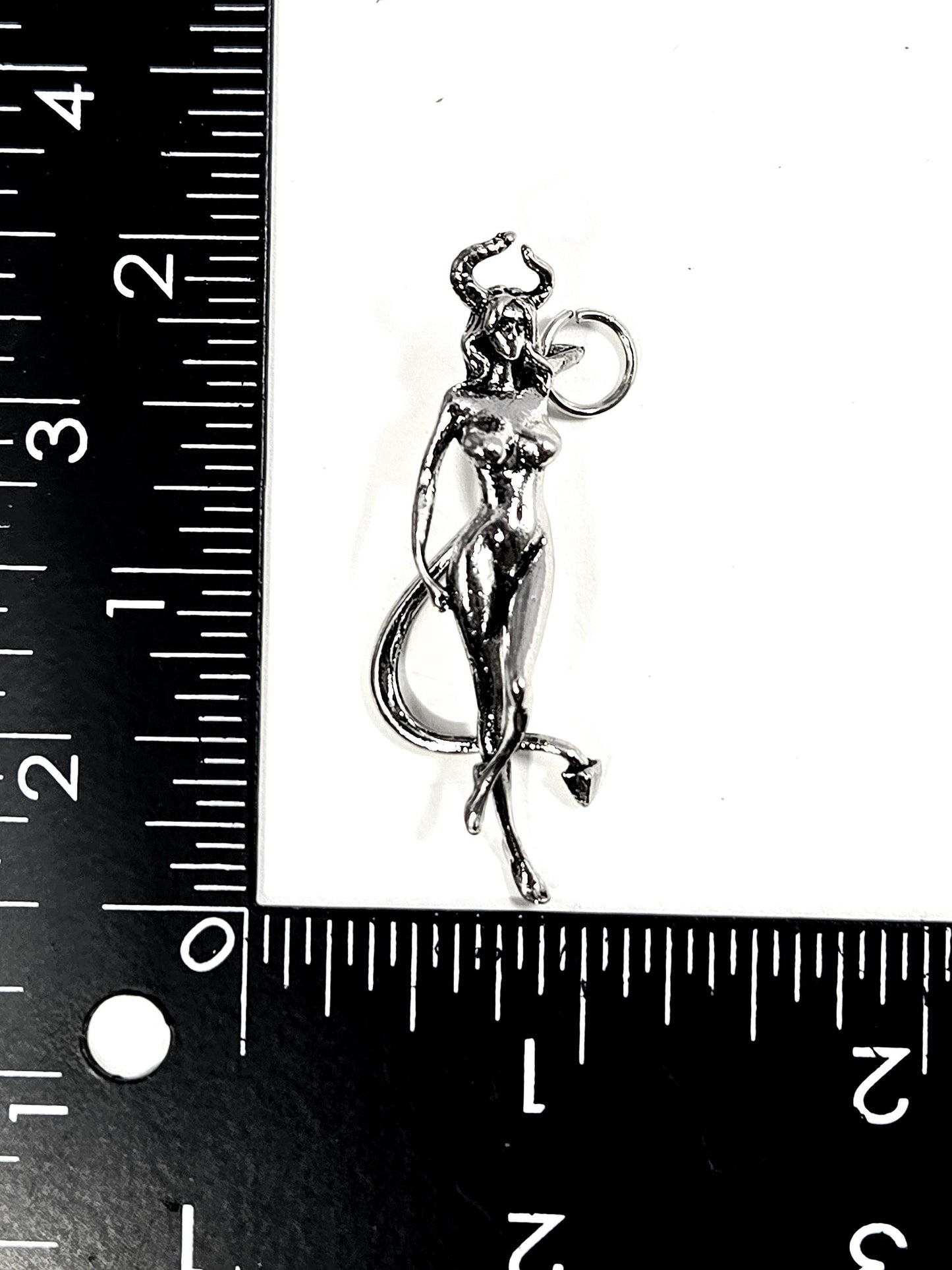 A silver-colored pendant in the shape of a female figure with horns and a tail, placed next to a ruler for size comparison.