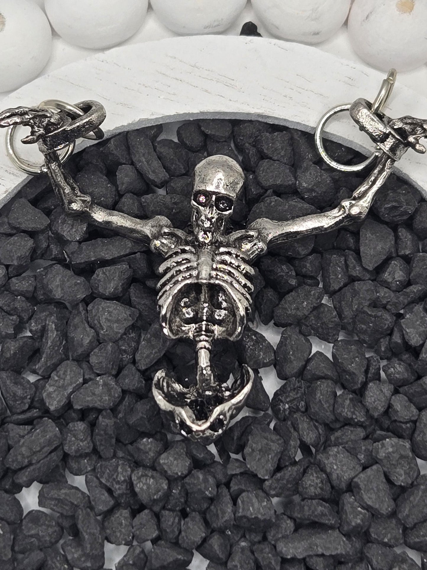 A silver skeleton necklace with a pendant is displayed on a bed of small black stones.