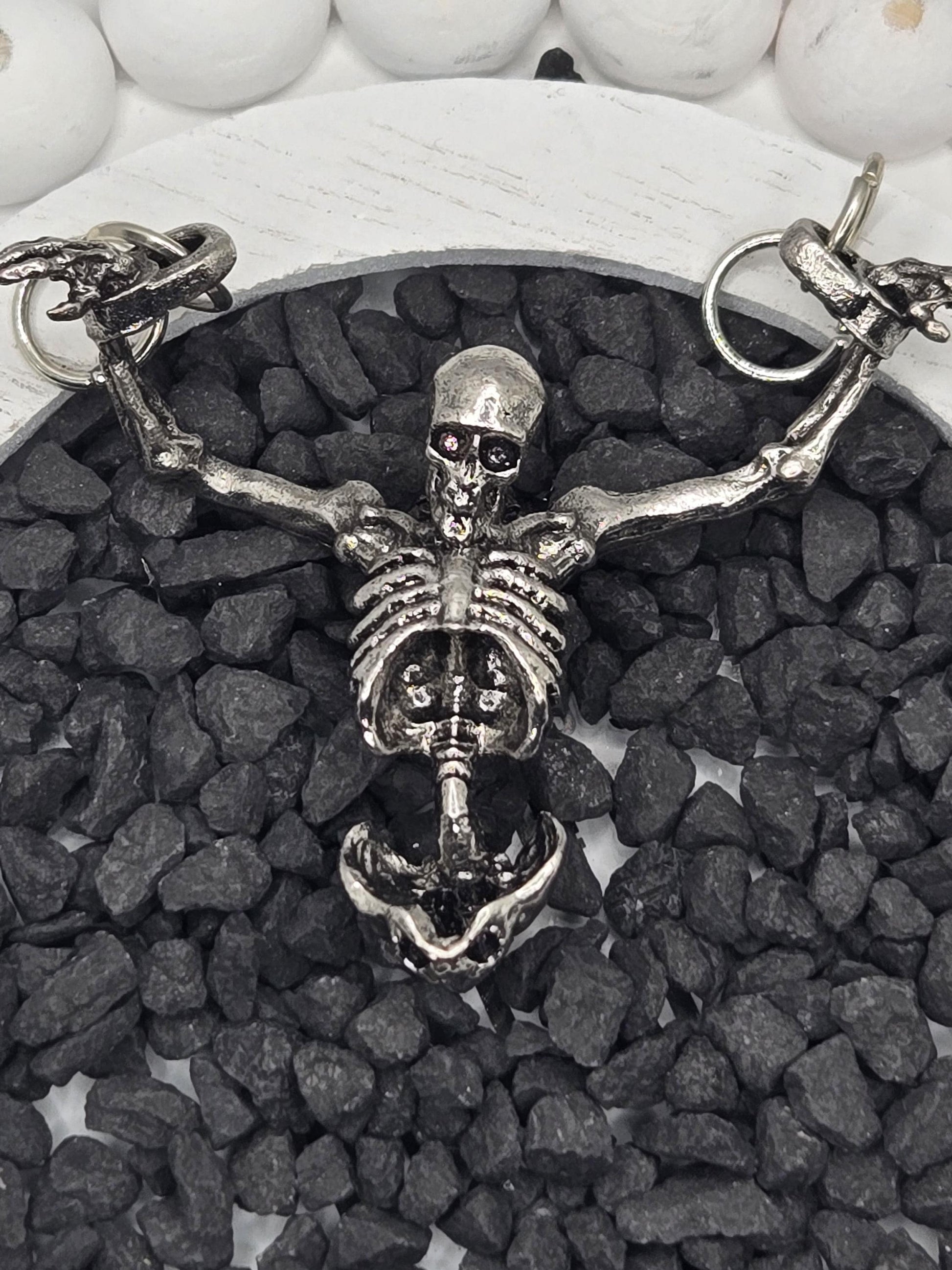 A silver skeleton necklace with a pendant is displayed on a bed of small black stones.