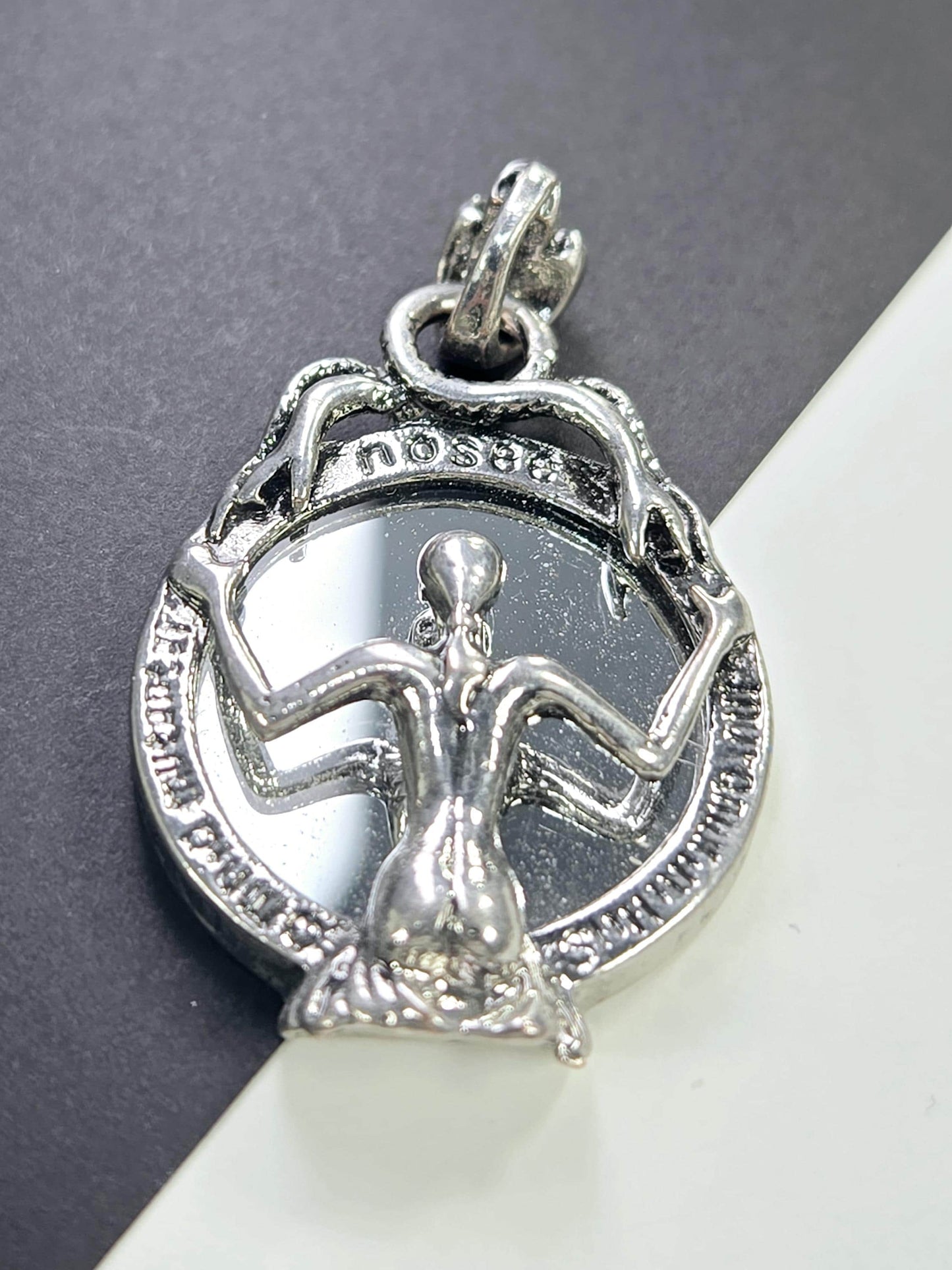 A silver-colored pendant or charm with an intricate design, featuring a figure or symbol within a circular frame.