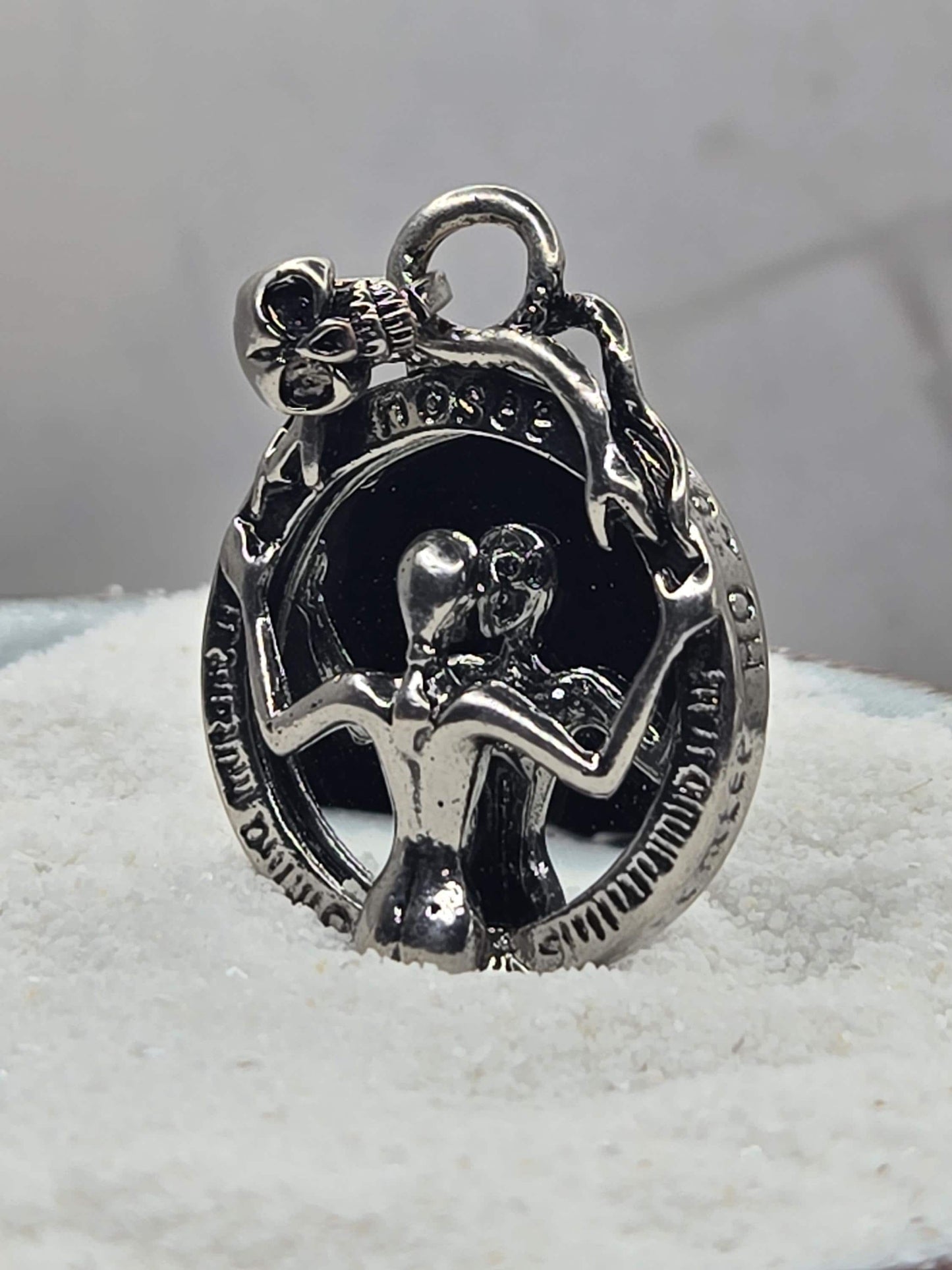 A silver-colored metal charm or pendant with an intricate design, including a skull and crossbones motif, and a small bell-like element.