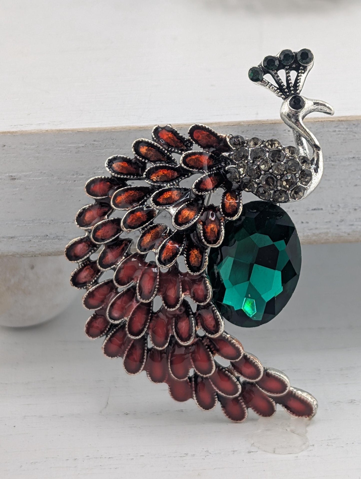 A decorative brooch in the shape of a peacock, with a large green gemstone on its back and a pattern of red and orange feathers on its tail.