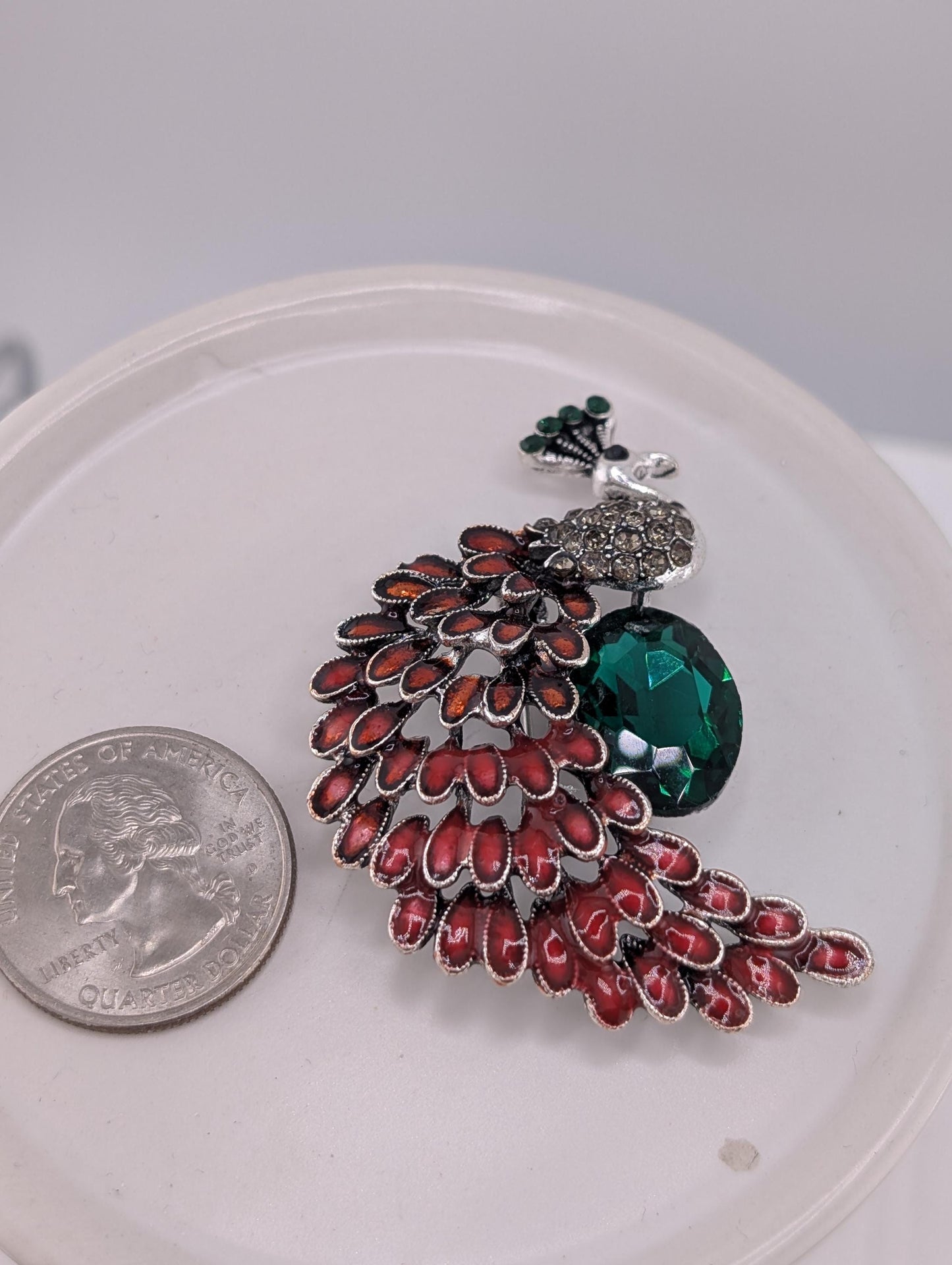 A decorative brooch with a peacock feather design, featuring a large green gemstone and red gemstones, is placed on a white plate next to a US quarter.
