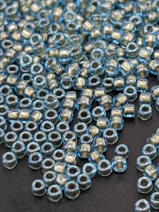 a close-up view of numerous small, clear beads scattered across a dark surface. The beads appear to be made of a transparent material, possibly glass or plastic, and are in various shades of blue and silver. The arrangement of the beads creates a visually interesting and textured composition.