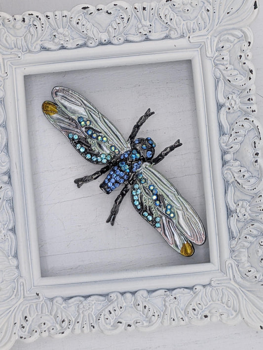 A decorative frame containing a large, intricately designed blue and silver insect, possibly a dragonfly or butterfly, with numerous small jewels or gemstones adorning its wings and body.
