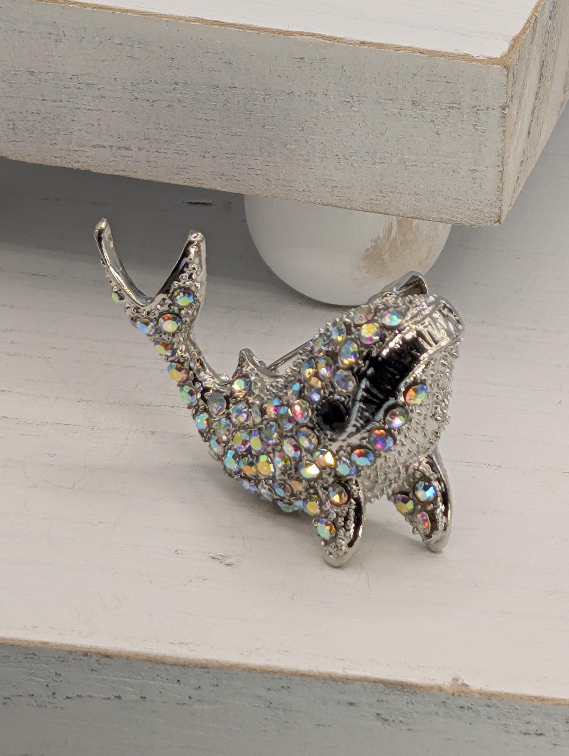 A silver-colored decorative fish brooch with a sparkling crystal design is displayed on a white surface.