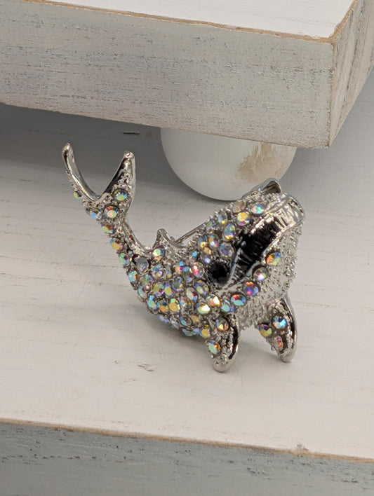 A silver-colored decorative fish brooch with a sparkling crystal design is displayed on a white surface.