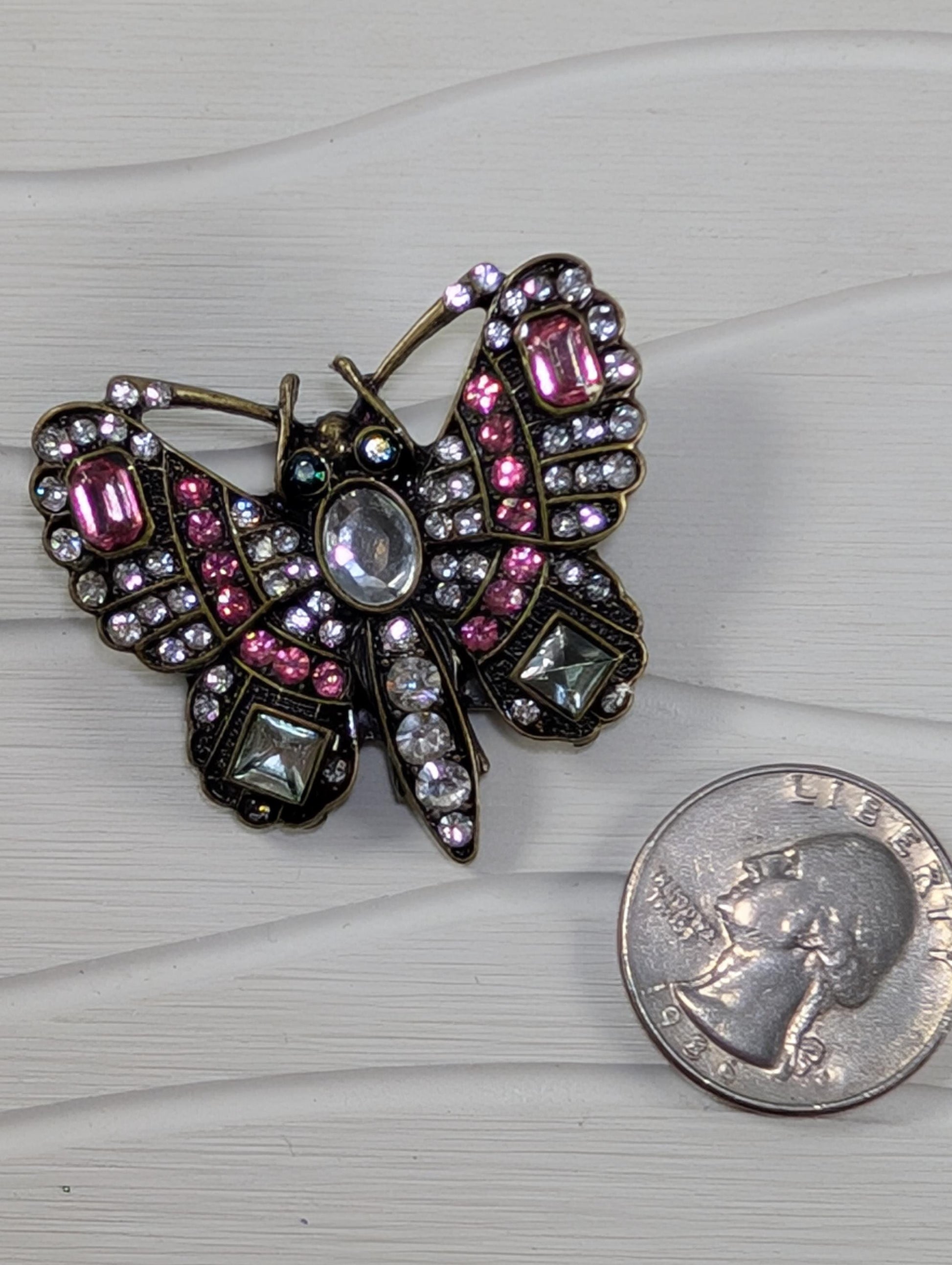 a decorative butterfly brooch adorned with pink and clear gemstones, placed next to a United States quarter.