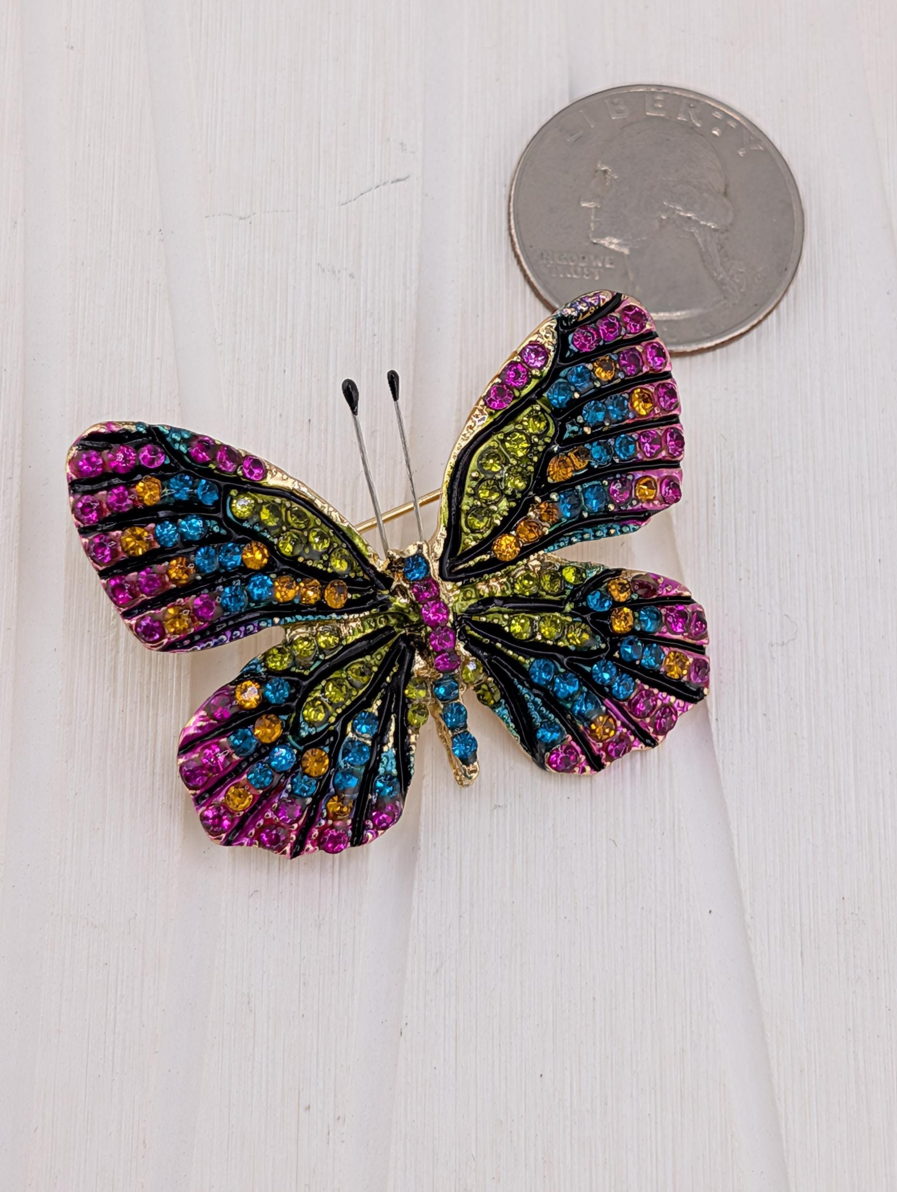 A colorful butterfly brooch with multicolored gemstones is placed on a white surface next to a coin.