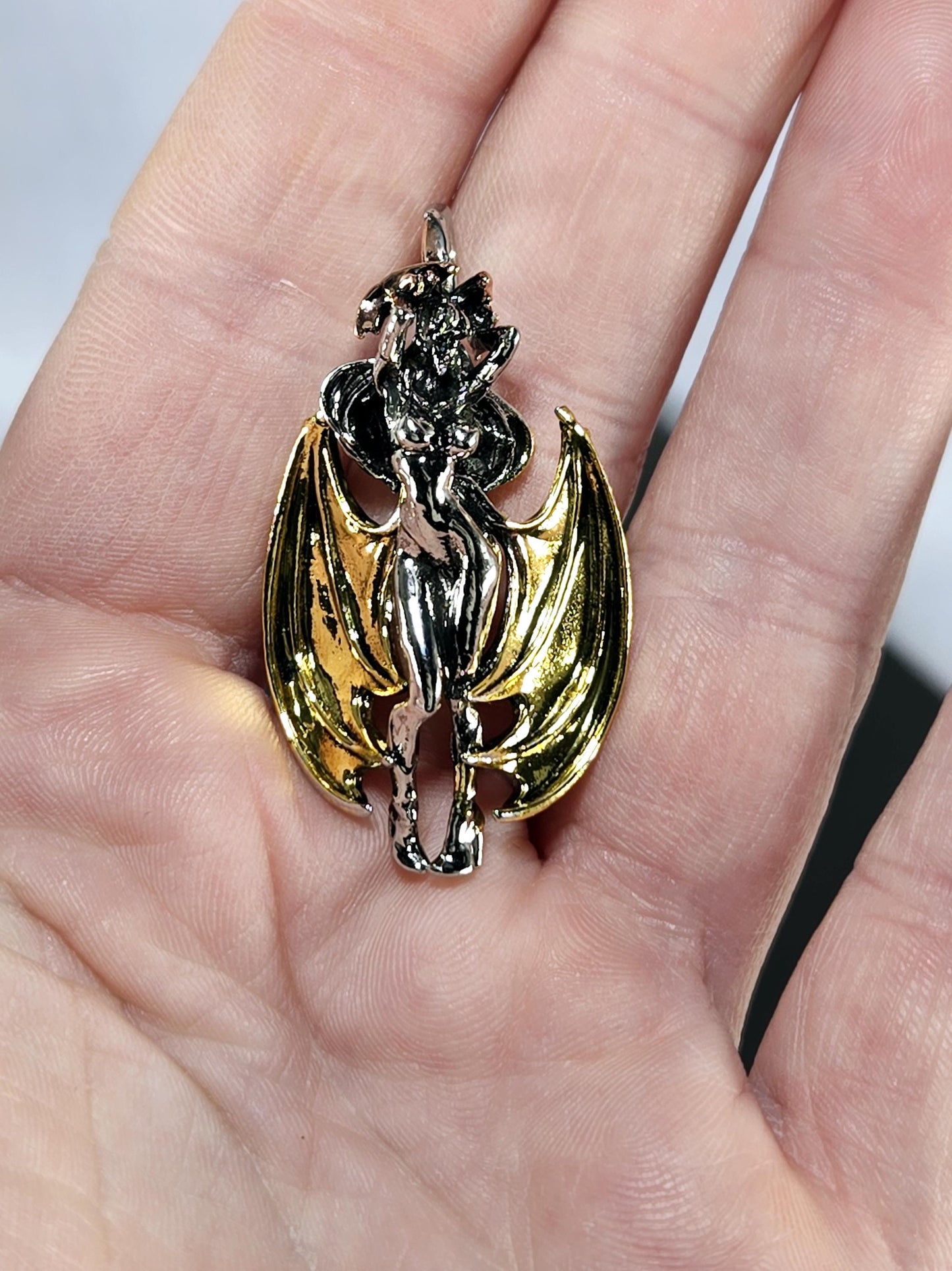A hand holding a decorative piece of jewelry, which appears to be a combination of a winged figure and a metallic design.