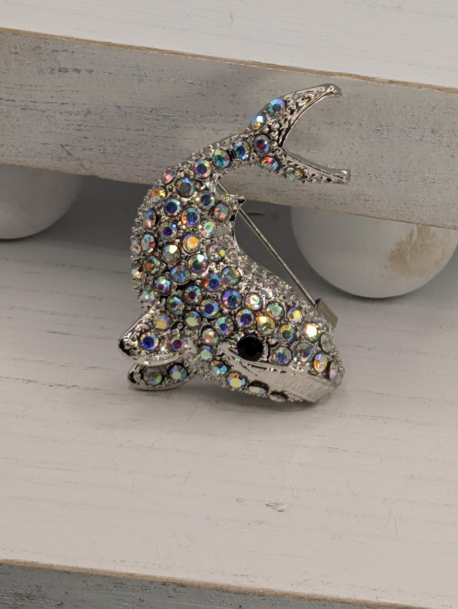 A silver brooch in the shape of a fish, adorned with numerous colorful rhinestones or crystals, is displayed on a white surface.