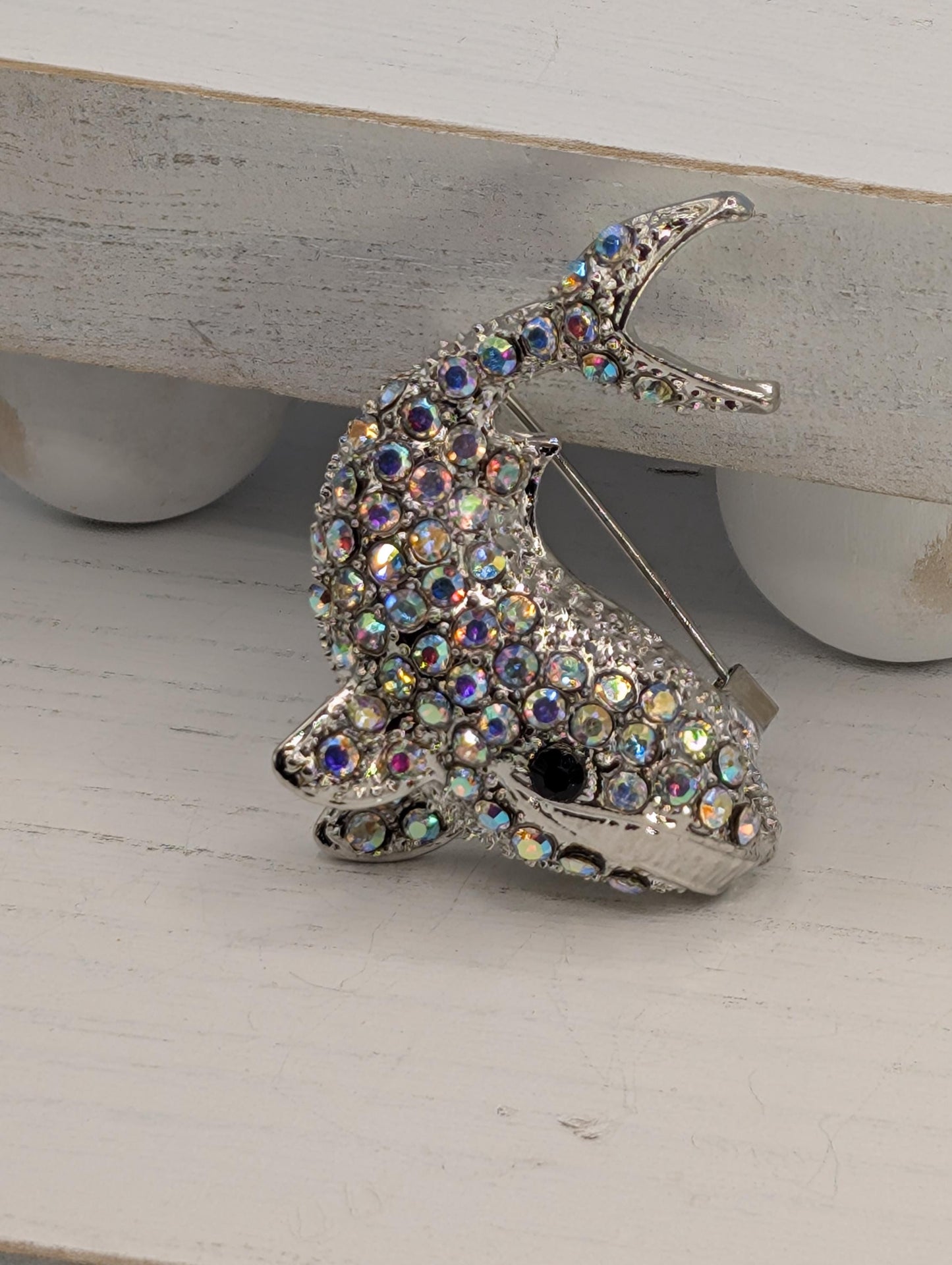 A silver fish brooch adorned with multicolored gemstones is displayed on a white surface.