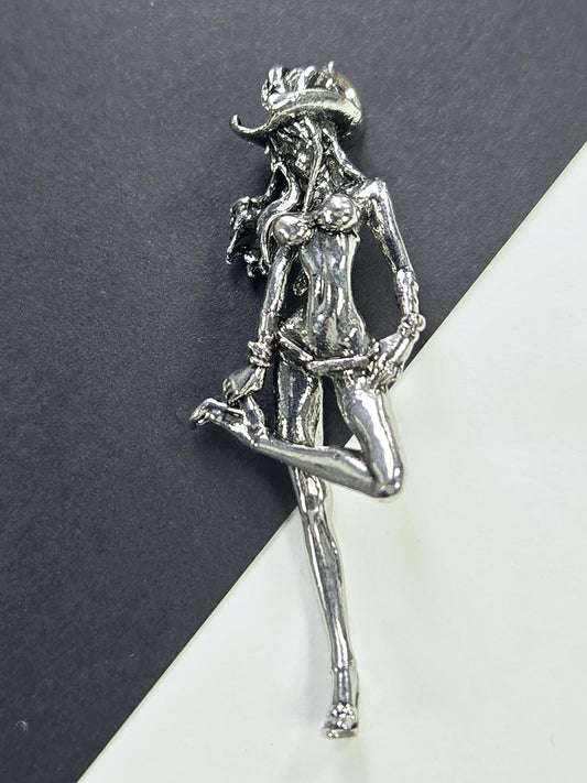 A silver metal brooch in the shape of a woman is shown on a black surface.