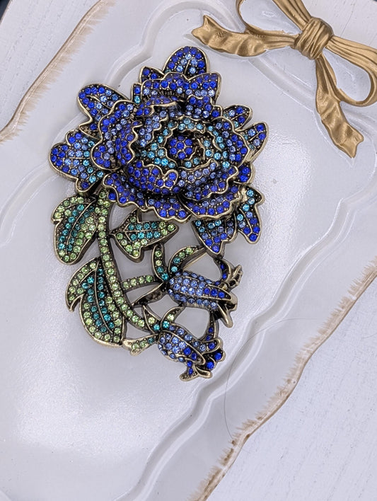 A decorative brooch with a large blue flower and green leaves, adorned with sparkling rhinestones.