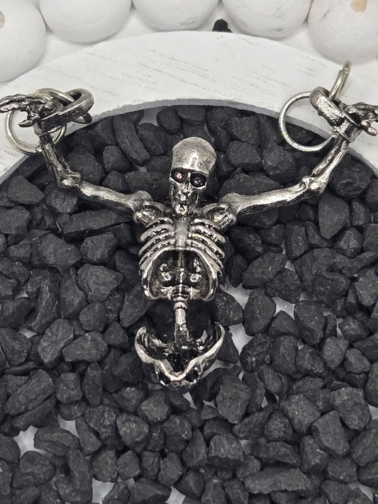 A silver skeleton necklace with a pendant is displayed on a bed of small black stones.