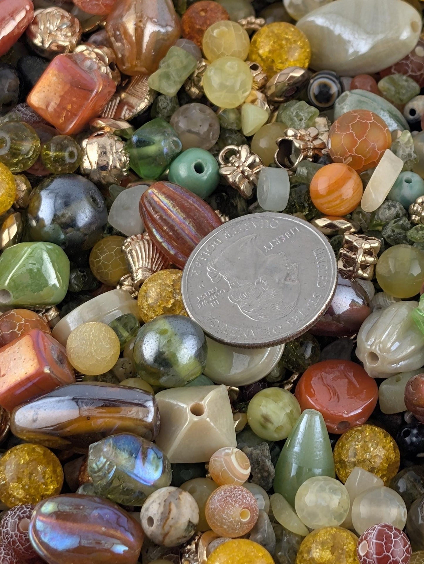 The image is a close-up of a pile of various colorful beads, including glass beads, metal beads, and other materials. There is a coin placed on top of the pile, which is a U.S. quarter.