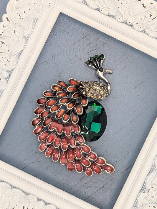 A decorative brooch with a large green gemstone and a cluster of red and orange gemstones, displayed in a white ornate frame.