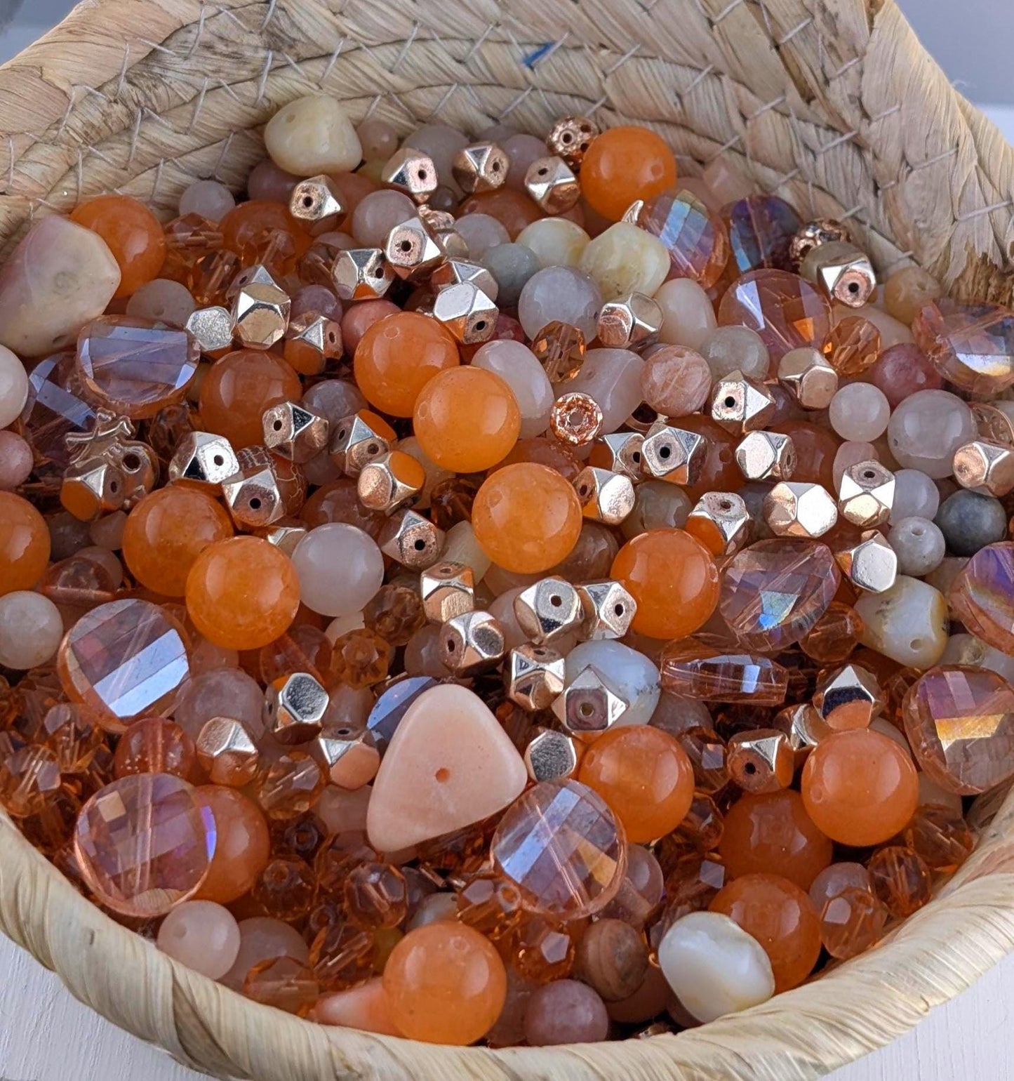 A woven basket filled with a variety of colorful beads, including clear, multicolored glass beads and metal beads in shades of orange, white, and silver.